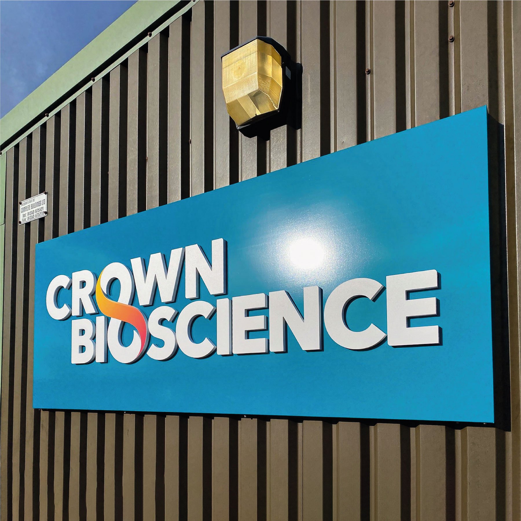 A light blue sign panel on a corrugated metal wall. The sign has white lettering saying 'Crown Bioscience' that is slightly 3D, the letter O in crown and bioscience create an infinity symbol together with a red to yellow gradient.