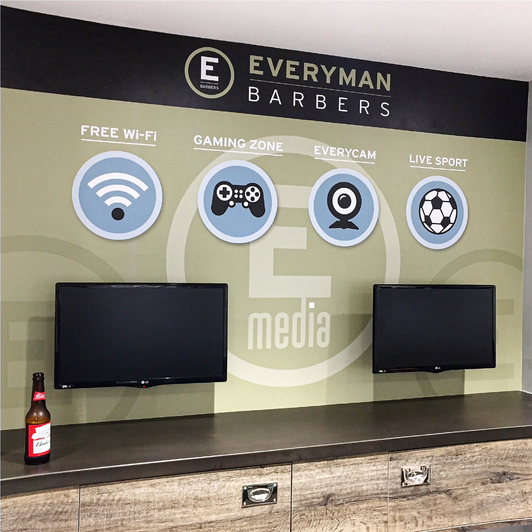 A Wall covering displaying Everyman Barbers Logo with a sage green background and symbols displaying free wifi, gaming zone, everycam and live sport. There are two monitors on the wall below the icons.