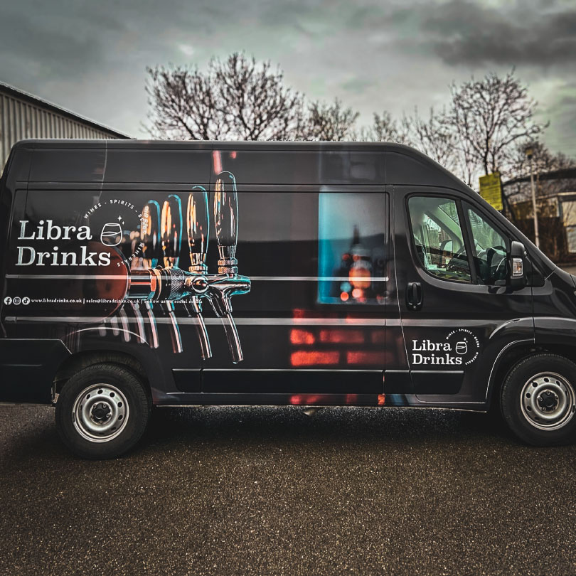 The image shows a van which has had a full cusotm printed wrap. The print on the side shows a photo of a series of beer taps infront of a blurred out bar with the Libra drinks logo and contact information over the top on the panel and door. The photo fades to black on the edges of the van to allow for a seamless blend to the solid black wrap on the front and rear of the van.