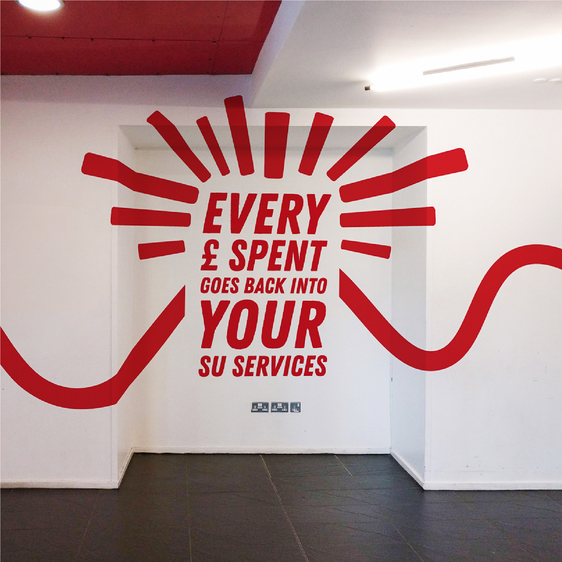 Written in red on a white wall 'Every pound Spent goes back into your SU Services' with red lines in an arch over the top.