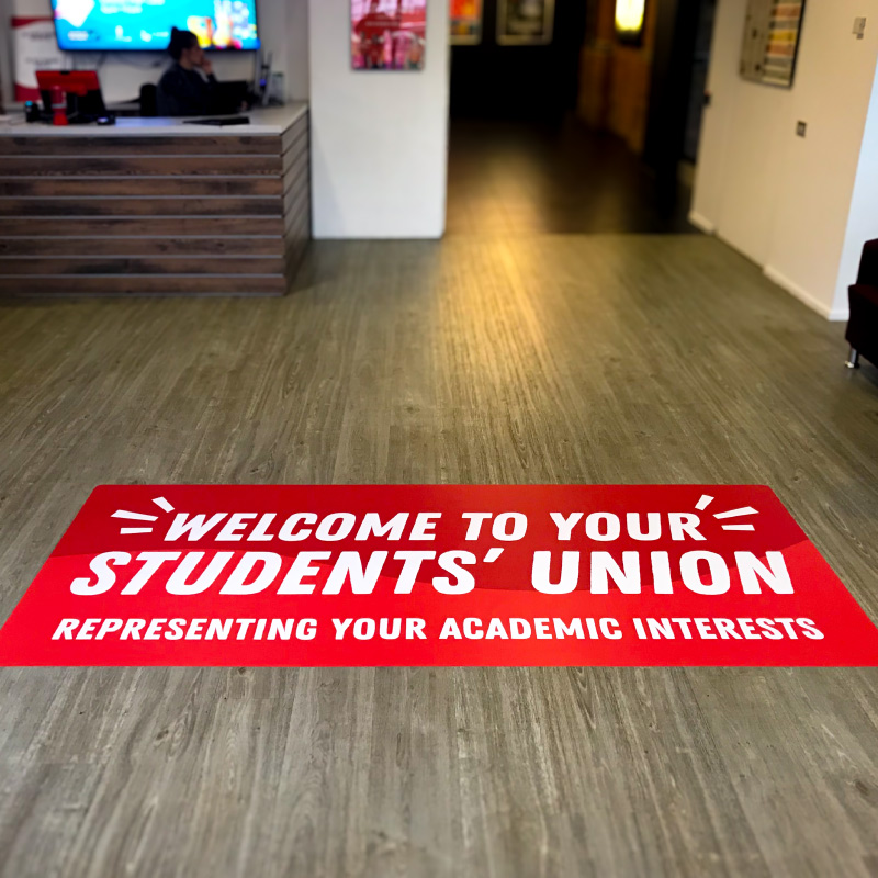 A wooden floor with a red rectangle graphic stuck to it. The graphic welcomes you to the students union.