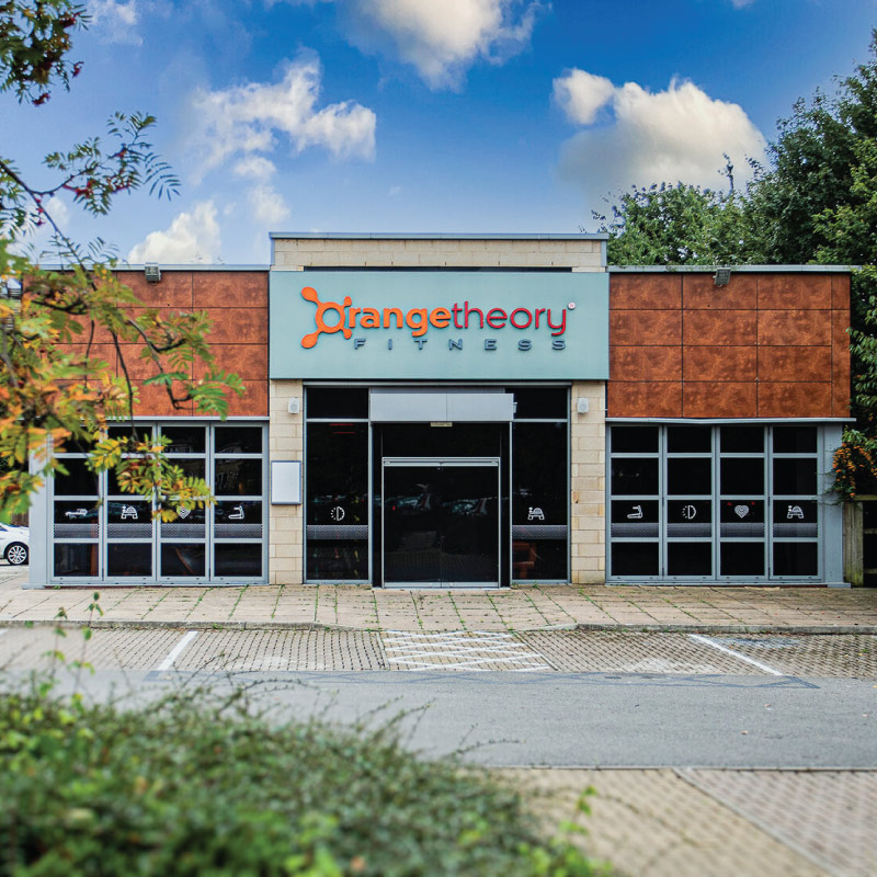The image is taken infront of a gym. The bottom half of the building has tall windows featuring white fitness icons. The top half of the gym has copper panels with a large grey sign panel in the middle. On the grey  sign panel is an illuminated 3D lettering sign saying 'Orange theory Fitness'. There are also white dots with fitness icons halfway down the windows.