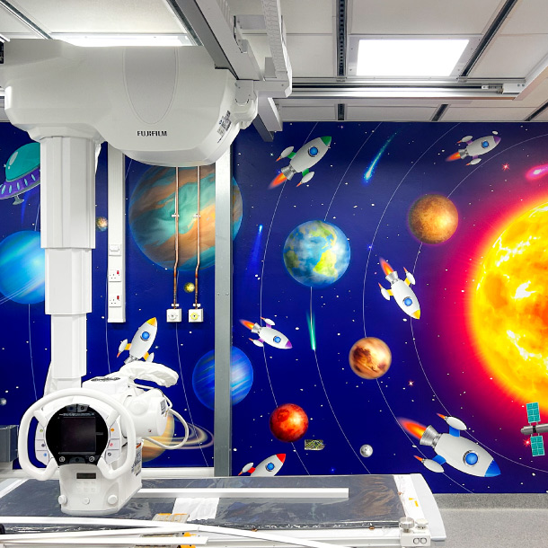 The image shows a hospital room with printed wall wrap. The wall wraps shows a space scene with the sun at one end, the planets are in orbit around it with some space ships and UFOs