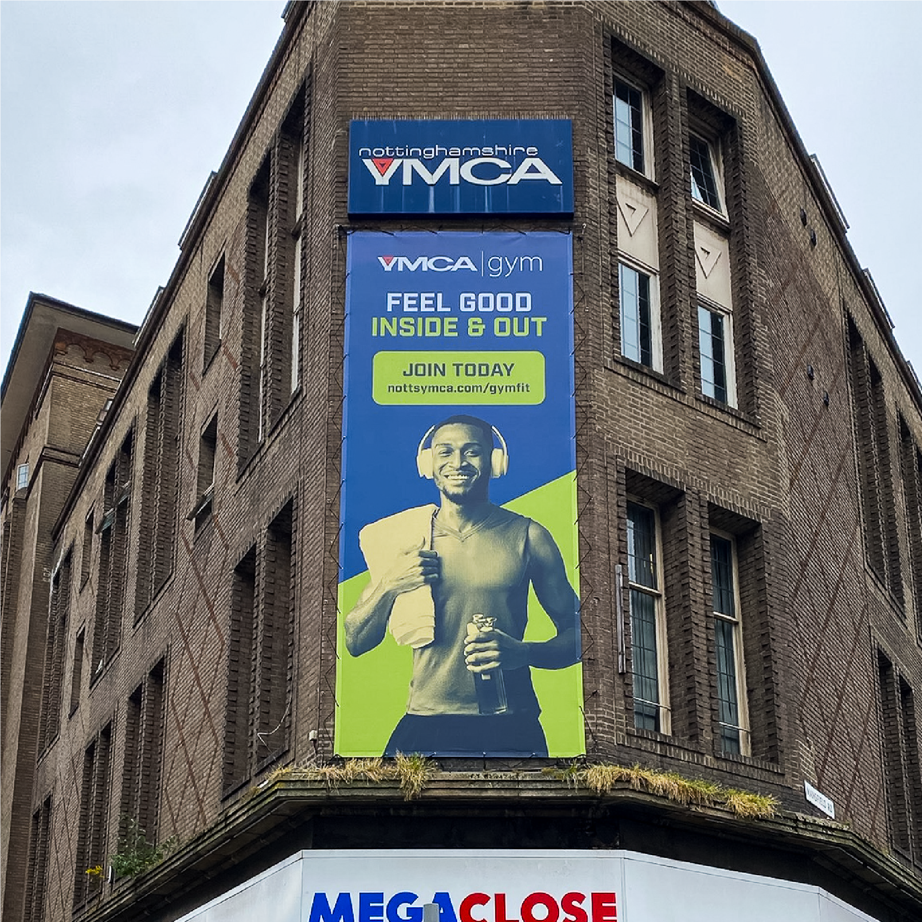 A large scale vertical banner on the corner of a brick building. The banner says 'Feel good inside and out, join today' with a blue background and green accents.