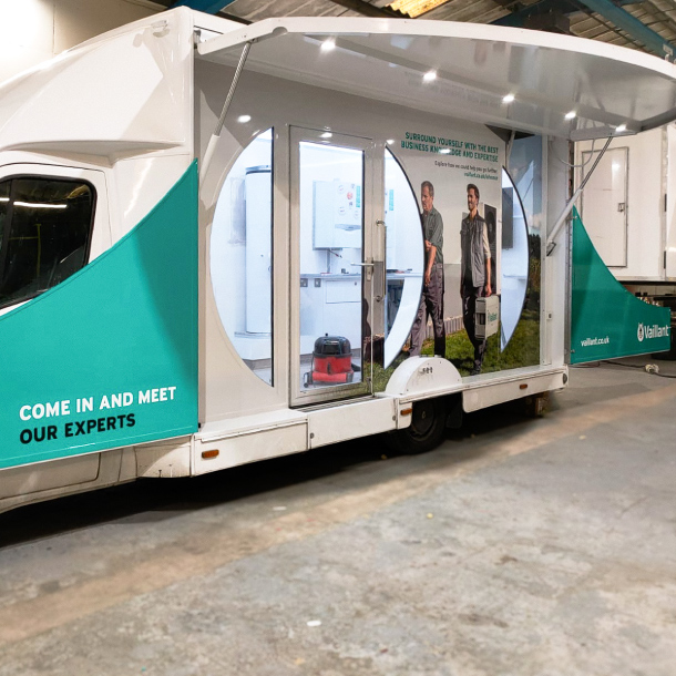 The image is of a Vaillant Show Vehicle. Two panels swing from the centre of the show vehicle to create outward facing information panels, these have been wrapped in a printed teal wrap as [per Vaillants brand colours. The Centre of the now opened up show vehicle has two engineers holding a Vaillant unit. Inside the vehicle are some boiler units for viewing. 